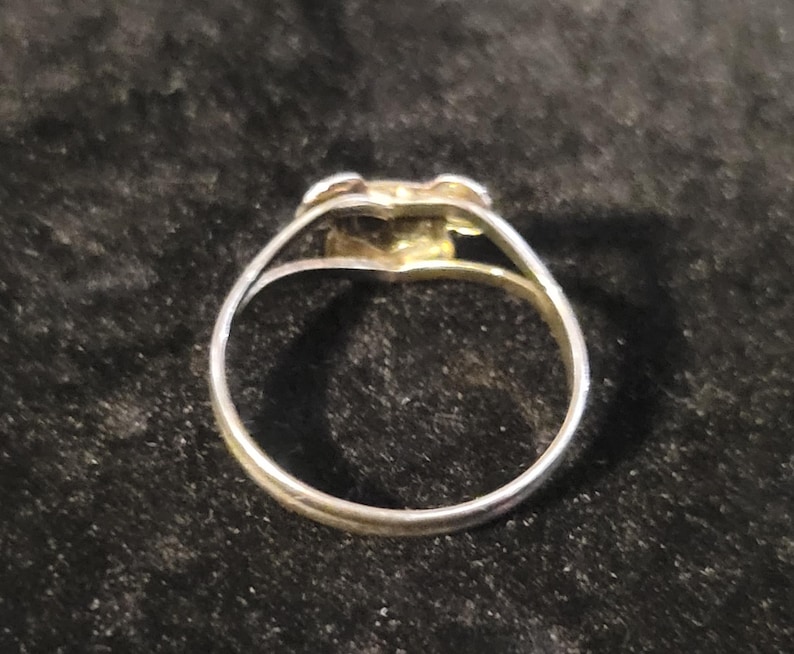 Puede incluir: Un anillo de metal en tono plateado con un dise&ntilde;o de v&aacute;stago dividido que converge en un elemento decorativo central. La banda es delgada y circular, mostrando un acabado met&aacute;lico pulido que refleja la luz en su estructura.