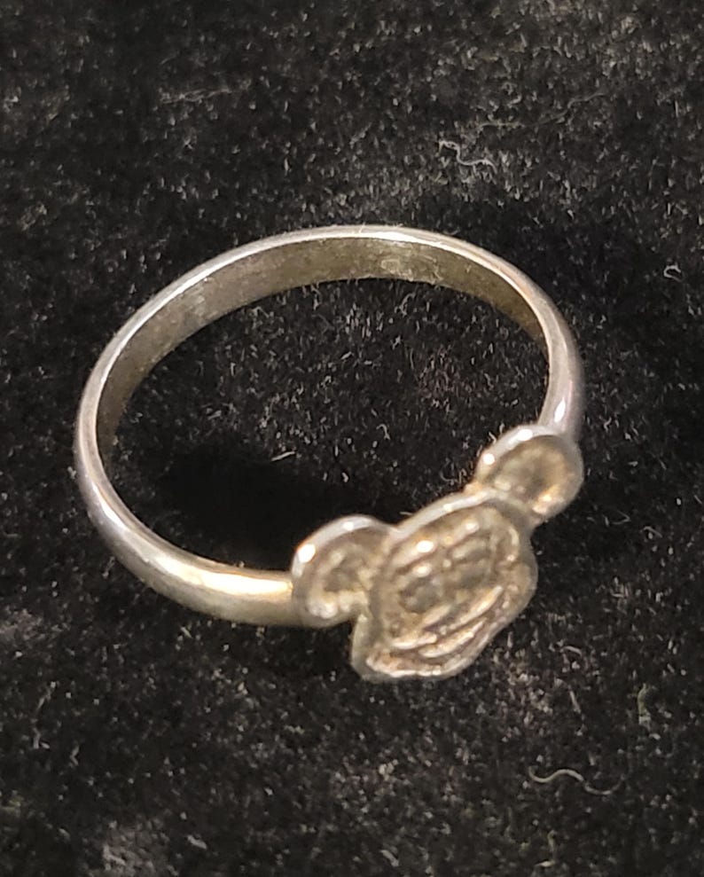 Pu&ograve; includere: Un anello color argento caratterizzato da un design in rilievo raffigurante la testa di un personaggio con due orecchie arrotondate. La fascia &egrave; liscia e circolare, con una finitura metallica che riflette la luce su tutta la superficie.