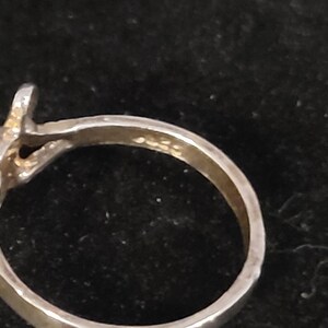 Puede incluir: Primer plano de un anillo plateado con dise&ntilde;o de nudo. El anillo tiene una superficie texturizada y un peque&ntilde;o elemento elevado en la parte superior. La banda es lisa y pulida. El anillo est&aacute; sobre un fondo oscuro.