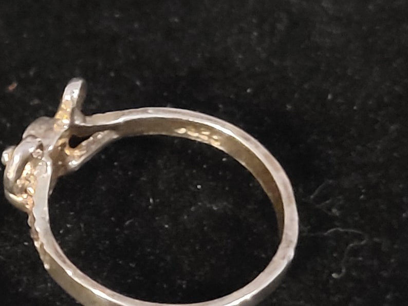 Puede incluir: Primer plano de un anillo plateado con dise&ntilde;o de nudo. El anillo tiene una superficie texturizada y un peque&ntilde;o elemento elevado en la parte superior. La banda es lisa y pulida. El anillo est&aacute; sobre un fondo oscuro.