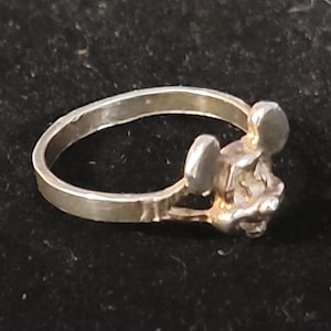 Puede incluir: Un anillo plateado con un dise&ntilde;o de rat&oacute;n estilizado. El anillo tiene una banda simple y una cabeza de rat&oacute;n detallada con orejas redondeadas. El anillo probablemente est&aacute; hecho de metal y es adecuado para el uso diario.