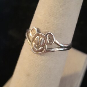 Puede incluir: Un anillo de tono plateado que presenta un motivo central con forma de cabeza de rat&oacute;n de dibujos animados con dos orejas redondeadas y una cara sonriente. La banda del anillo se divide en dos l&iacute;neas finas y paralelas en el dise&ntilde;o central.