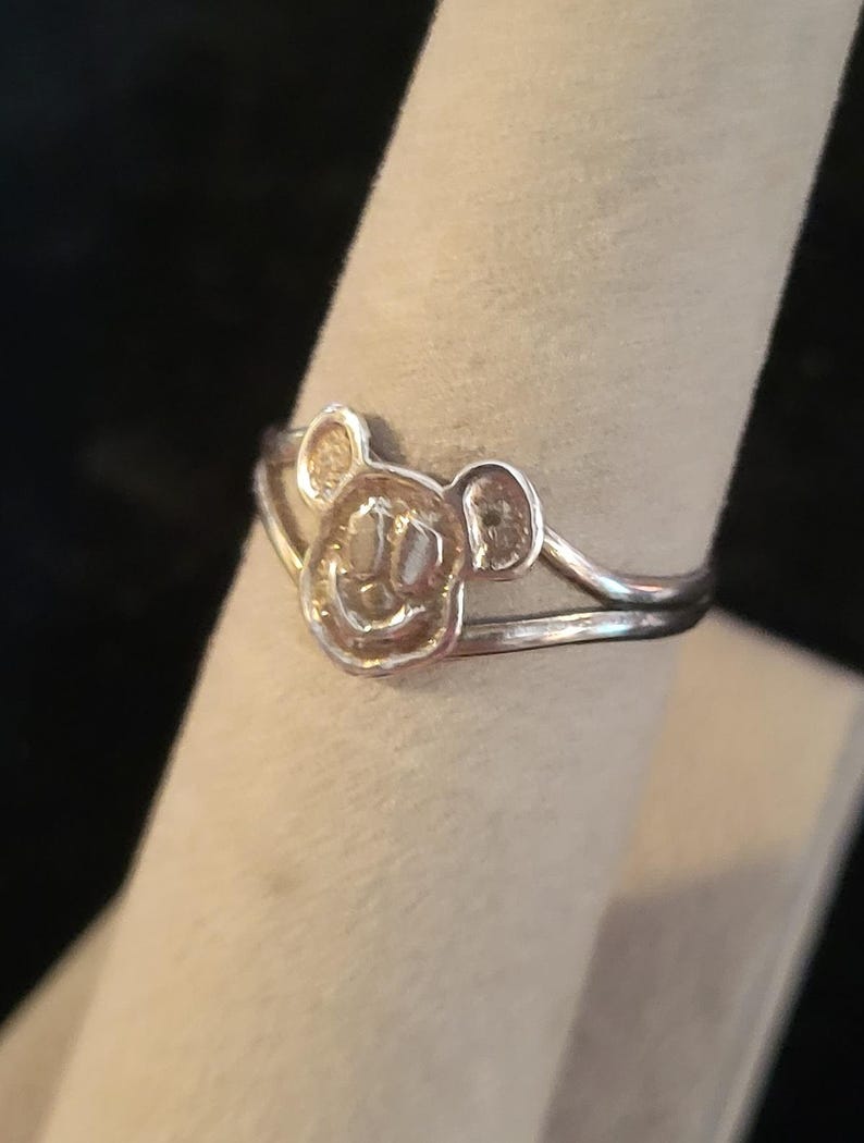 Puede incluir: Un anillo de tono plateado que presenta un motivo central con forma de cabeza de rat&oacute;n de dibujos animados con dos orejas redondeadas y una cara sonriente. La banda del anillo se divide en dos l&iacute;neas finas y paralelas en el dise&ntilde;o central.