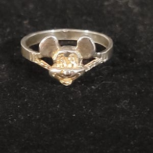 Puede incluir: Un anillo de plata con un dise&ntilde;o de Mickey Mouse. El anillo presenta una cara detallada de Mickey Mouse con detalles dorados. El anillo est&aacute; sobre un fondo oscuro.