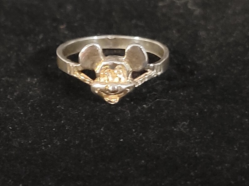 Puede incluir: Un anillo de plata con un dise&ntilde;o de Mickey Mouse. El anillo presenta una cara detallada de Mickey Mouse con detalles dorados. El anillo est&aacute; sobre un fondo oscuro.