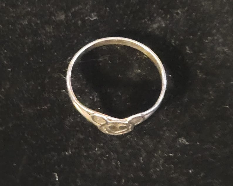 Puede incluir: Un anillo de metal plateado que presenta un elemento de dise&ntilde;o central con forma de cabeza de rat&oacute;n estilizada, con dos orejas circulares y una cara redondeada. La banda es delgada y circular, manteniendo un acabado met&aacute;lico uniforme.