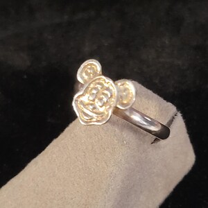 Pu&ograve; includere: Un anello in metallo color argento caratterizzato dal volto scolpito e in rilievo di un topo dei cartoni animati con grandi orecchie arrotondate. L'anello presenta una fascia liscia e lucida e un design frontale dettagliato e strutturato.