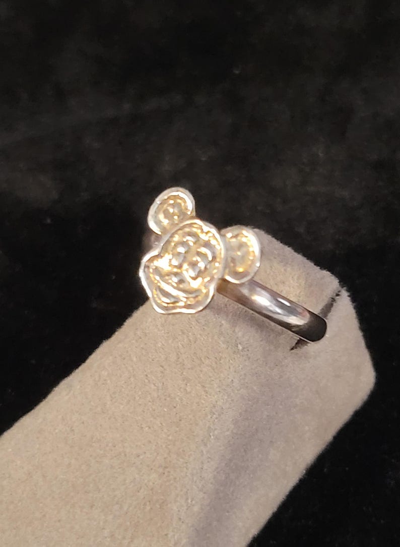 Pu&ograve; includere: Un anello in metallo color argento caratterizzato dal volto scolpito e in rilievo di un topo dei cartoni animati con grandi orecchie arrotondate. L'anello presenta una fascia liscia e lucida e un design frontale dettagliato e strutturato.