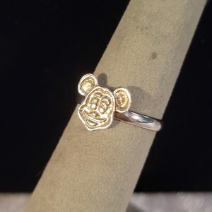 Pu&ograve; includere: Un anello color argento con un emblema in metallo color oro in rilievo raffigurante una testa di topo dei cartoni animati con orecchie arrotondate e un viso sorridente. La fascia dell'anello &egrave; liscia e lucida, in contrasto con il viso dettagliato.