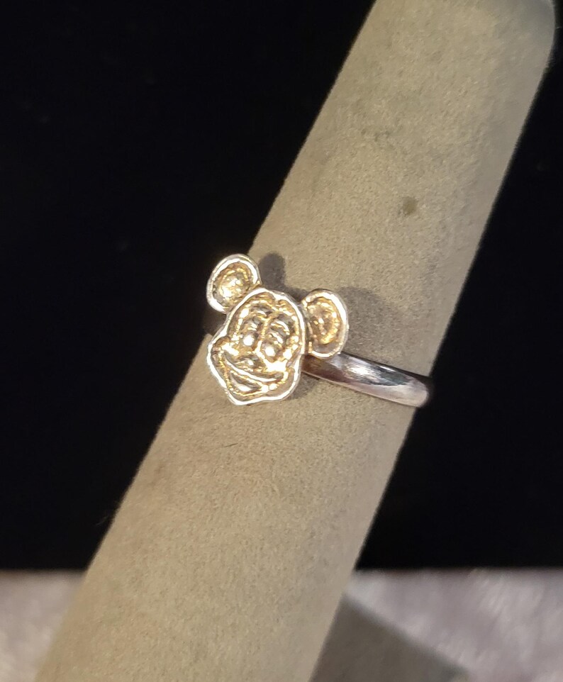 Pu&ograve; includere: Un anello color argento con un emblema in metallo color oro in rilievo raffigurante una testa di topo dei cartoni animati con orecchie arrotondate e un viso sorridente. La fascia dell'anello &egrave; liscia e lucida, in contrasto con il viso dettagliato.