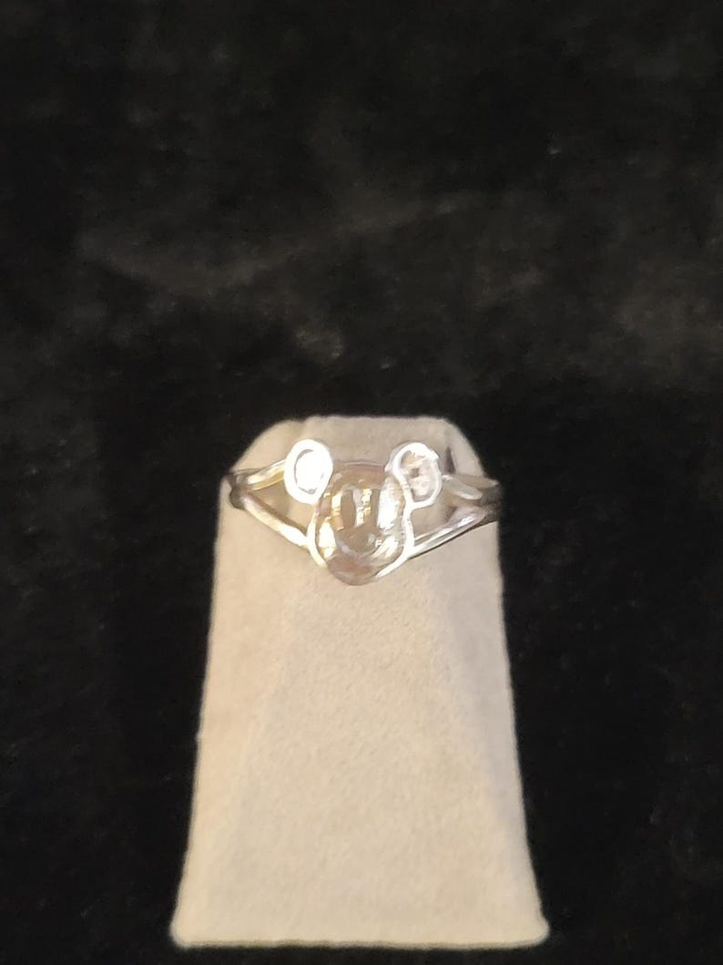 Puede incluir: Un anillo de tono plateado que presenta un icono central con forma de cabeza y dos orejas redondeadas. La banda del anillo se divide en dos segmentos met&aacute;licos finos y pulidos que se conectan a los lados del dise&ntilde;o central inspirado en un personaje.