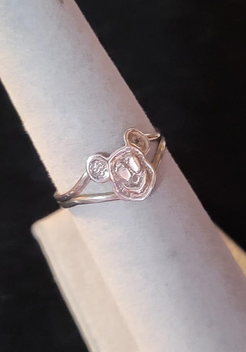 Puede incluir: Un anillo de metal plateado que presenta un dise&ntilde;o central recortado de una cabeza de rat&oacute;n de dibujos animados con dos orejas redondeadas. La banda es delgada y lisa, conect&aacute;ndose a la base del motivo de la cabeza del rat&oacute;n.