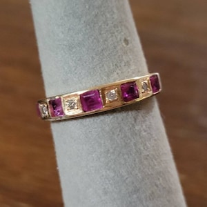May include: A gold band ring with a row of pink square-shaped gemstones and smaller clear round gemstones.