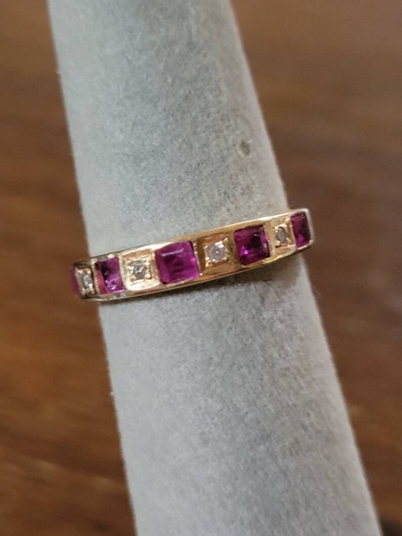 May include: A gold band ring with a row of pink square-shaped gemstones and smaller clear round gemstones.