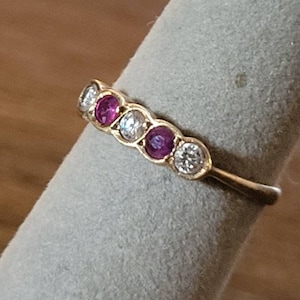 May include: A gold band ring with alternating pink and white gemstones. The ring is set on a white velvet display stand.
