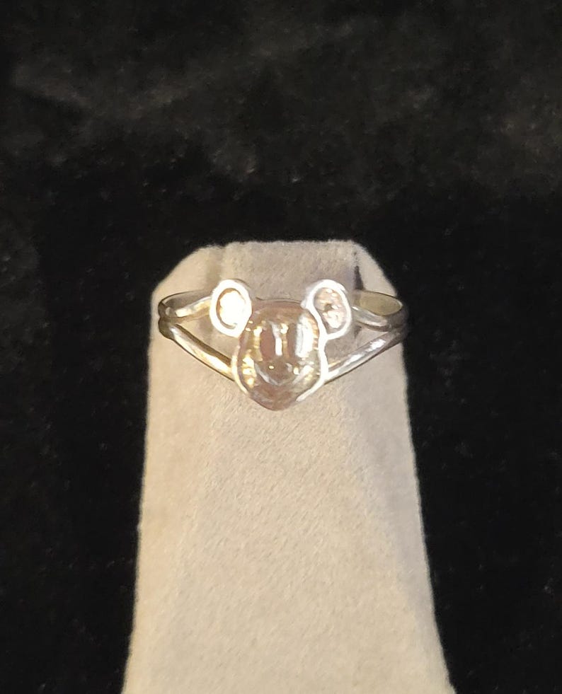 Puede incluir: Un anillo de tono plateado que presenta un motivo central con forma de cabeza de rat&oacute;n con dos orejas circulares. La banda se divide en dos secciones delgadas que se unen en la base de la cabeza, creando un dise&ntilde;o sim&eacute;trico con acabado pulido.
