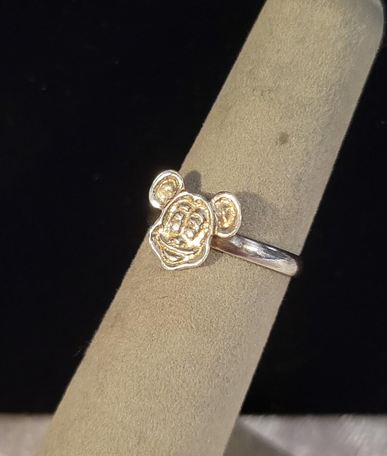 Pu&ograve; includere: Un anello in metallo color argento con un emblema in rilievo che raffigura la testa di Topolino sulla parte anteriore. La fascia &egrave; liscia e lucida, mentre il volto del personaggio presenta dettagli incisi con una finitura metallica uniforme.