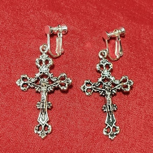 May include: A pair of silver clip-on earrings featuring ornate cross designs with a figure of Jesus Christ on each cross. The crosses have intricate details and a slightly aged finish.
