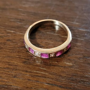 May include: A gold band ring with a row of pink square-shaped stones and smaller clear stones set in the band.