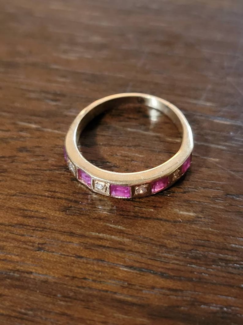 May include: A gold band ring with a row of pink square-shaped stones and smaller clear stones set in the band.