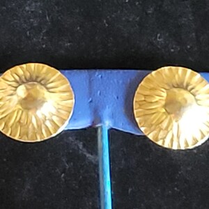 May include: A pair of gold-toned earrings with a textured, sunburst design. The earrings are round and have a slightly raised center.