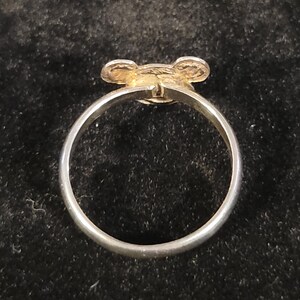 Pu&ograve; includere: Un anello color argento con un piccolo ornamento circolare piatto sulla parte superiore. L'ornamento presenta due sporgenze arrotondate simili a orecchie sui lati, creando una silhouette che ricorda una testa di animale stilizzata o un topo.