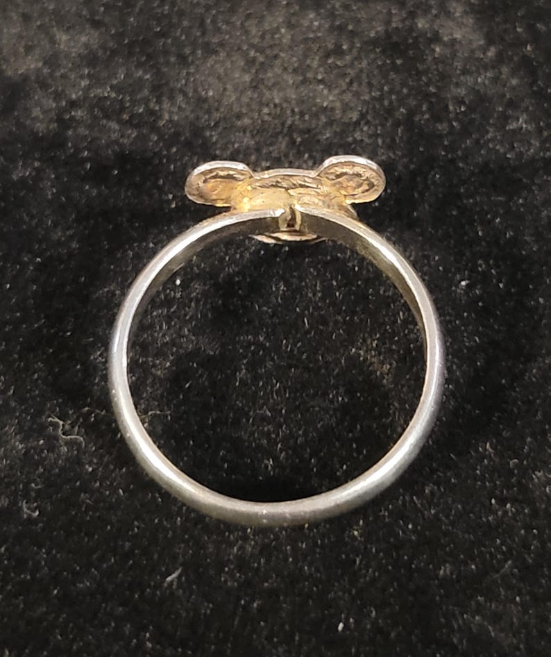 Pu&ograve; includere: Un anello color argento con un piccolo ornamento circolare piatto sulla parte superiore. L'ornamento presenta due sporgenze arrotondate simili a orecchie sui lati, creando una silhouette che ricorda una testa di animale stilizzata o un topo.