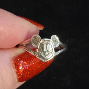 Puede incluir: Un anillo de metal plateado que presenta un dise&ntilde;o de cabeza de rat&oacute;n estilizada con orejas redondeadas y una cara sonriente. La banda del anillo se divide en dos secciones delgadas al conectarse con el motivo central de la cabeza del rat&oacute;n.