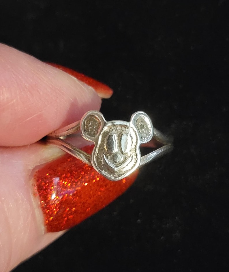 Puede incluir: Un anillo de metal plateado que presenta un dise&ntilde;o de cabeza de rat&oacute;n estilizada con orejas redondeadas y una cara sonriente. La banda del anillo se divide en dos secciones delgadas al conectarse con el motivo central de la cabeza del rat&oacute;n.
