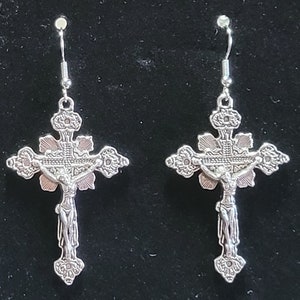 May include: A pair of silver dangle earrings featuring a detailed cross with a figure of Jesus Christ. The cross has a floral design and is suspended from a silver hook.