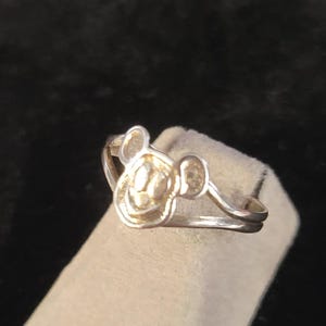 Puede incluir: Un anillo de tono plateado que presenta un motivo central con forma de cabeza de rat&oacute;n de dibujos animados con orejas redondeadas. La banda est&aacute; dividida en dos hilos plateados finos y paralelos que se conectan a los lados del dise&ntilde;o de la cabeza.