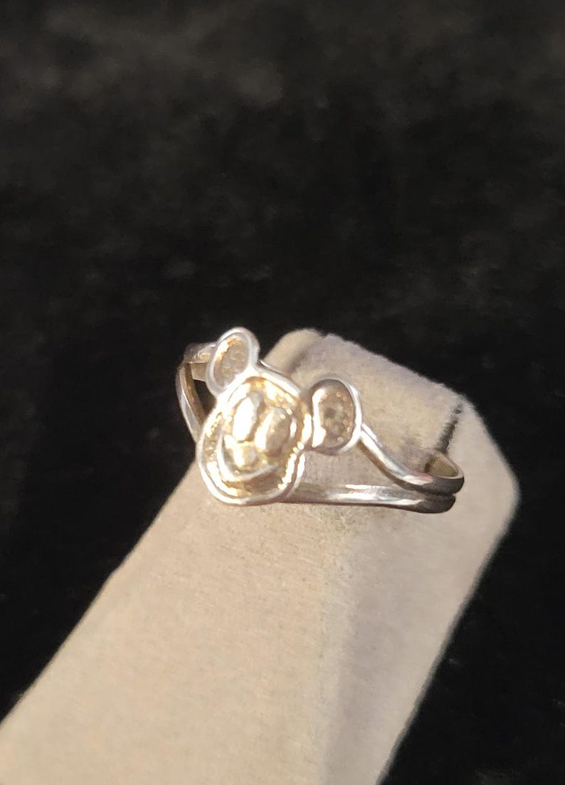 Puede incluir: Un anillo de tono plateado que presenta un motivo central con forma de cabeza de rat&oacute;n de dibujos animados con orejas redondeadas. La banda est&aacute; dividida en dos hilos plateados finos y paralelos que se conectan a los lados del dise&ntilde;o de la cabeza.