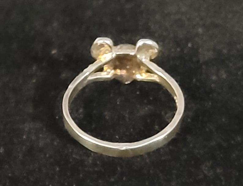 Puede incluir: Un anillo plateado con un peque&ntilde;o dise&ntilde;o en forma de oso de color dorado. El anillo tiene una banda simple y una cabeza de oso detallada con peque&ntilde;as orejas. El anillo est&aacute; sobre un fondo oscuro.