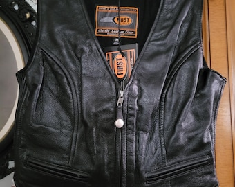 May include: Black leather vest with a V-neck, two front pockets, and a silver zipper. The vest has a tag that reads "FIRST Classic Leather Gear". The vest is hanging on a black hanger.
