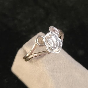 Puede incluir: Un anillo de tono plateado que presenta una cara circular elevada con el contorno en relieve de una cabeza de rat&oacute;n de dibujos animados. La banda es delgada y met&aacute;lica, con un dise&ntilde;o dividido que se conecta al emblema central.