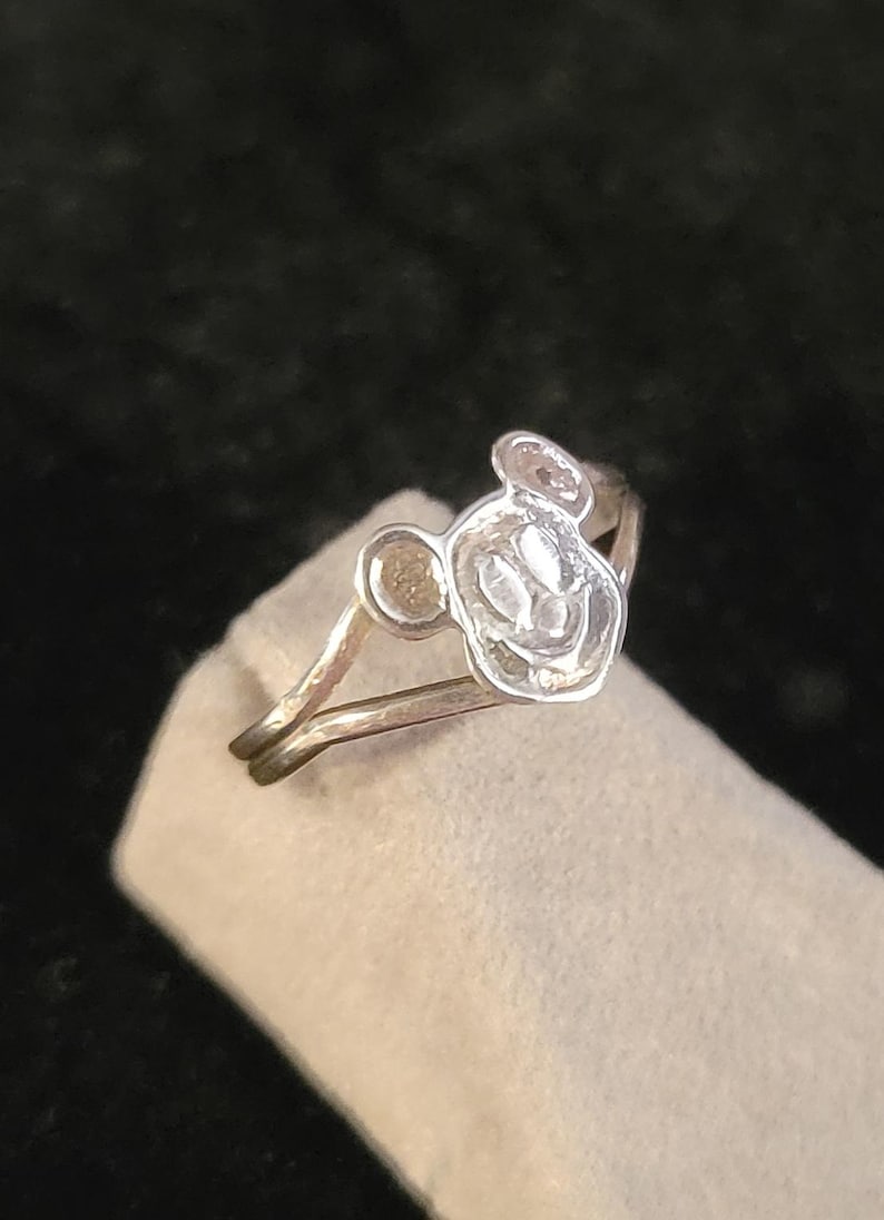 Puede incluir: Un anillo de tono plateado que presenta una cara circular elevada con el contorno en relieve de una cabeza de rat&oacute;n de dibujos animados. La banda es delgada y met&aacute;lica, con un dise&ntilde;o dividido que se conecta al emblema central.