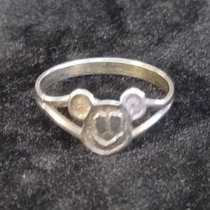Puede incluir: Un anillo de metal plateado con un motivo central en forma de cabeza de rat&oacute;n estilizada con dos orejas redondeadas y una cara circular. La banda del anillo es delgada y se divide en dos segmentos al conectar con el dise&ntilde;o central de la cabeza.