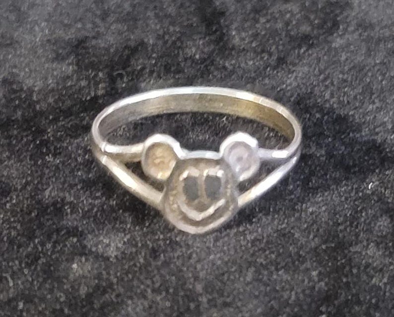 Puede incluir: Un anillo de metal plateado con un motivo central en forma de cabeza de rat&oacute;n estilizada con dos orejas redondeadas y una cara circular. La banda del anillo es delgada y se divide en dos segmentos al conectar con el dise&ntilde;o central de la cabeza.