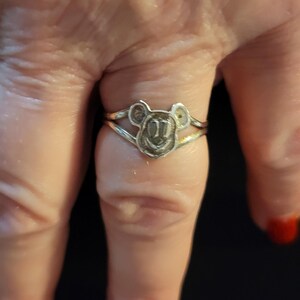 Puede incluir: Un anillo de metal plateado con un dise&ntilde;o central recortado en forma de cabeza de rat&oacute;n de dibujos animados con orejas redondeadas y cara sonriente. La banda del anillo se divide en dos l&iacute;neas finas y paralelas que se unen a la cabeza.