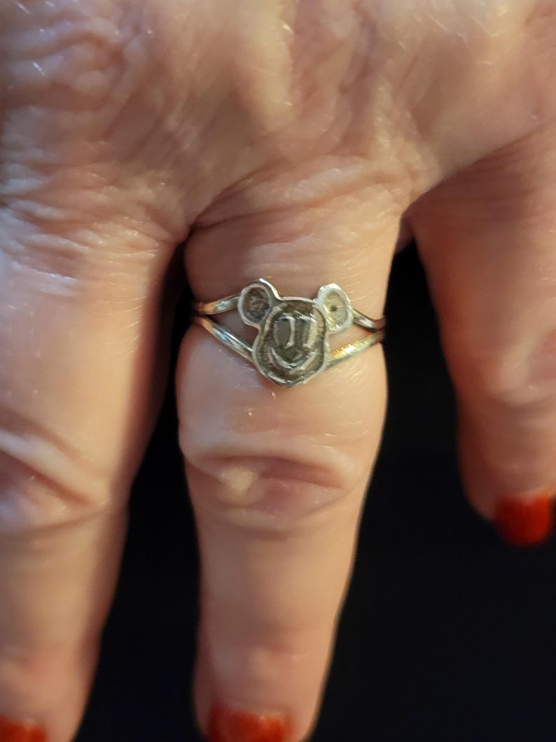 Puede incluir: Un anillo de metal plateado con un dise&ntilde;o central recortado en forma de cabeza de rat&oacute;n de dibujos animados con orejas redondeadas y cara sonriente. La banda del anillo se divide en dos l&iacute;neas finas y paralelas que se unen a la cabeza.