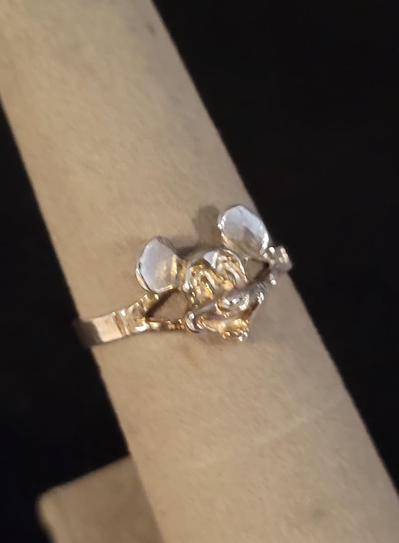 Puede incluir: Un anillo plateado con un dise&ntilde;o de rat&oacute;n estilizado. El anillo tiene una banda fina, y la cabeza y las orejas del rat&oacute;n son claramente visibles. El anillo probablemente est&aacute; hecho de una aleaci&oacute;n de metal y es adecuado para el uso diario.
