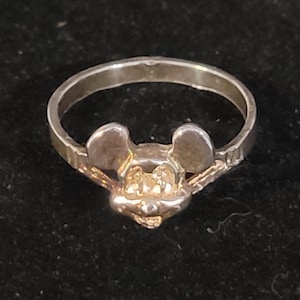 Puede incluir: Un anillo de plata con un dise&ntilde;o de Mickey Mouse. El anillo tiene una cara y orejas detalladas, con una banda lisa. El anillo probablemente est&aacute; hecho de plata de ley o un metal similar. El anillo es una joya.