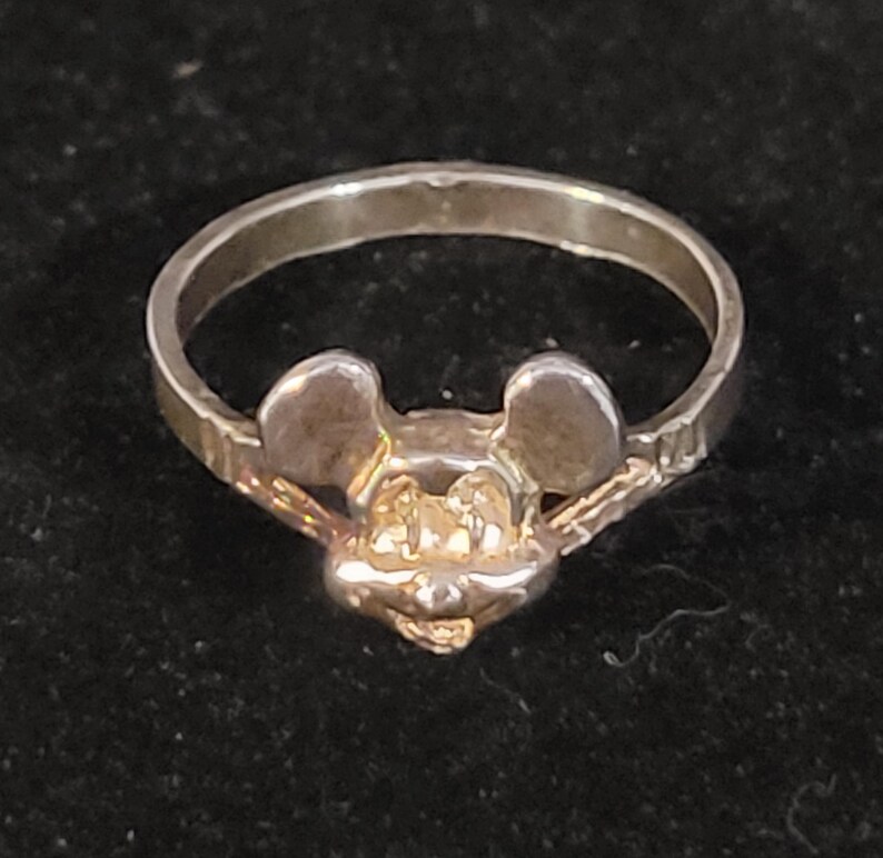 Puede incluir: Un anillo de plata con un dise&ntilde;o de Mickey Mouse. El anillo tiene una cara y orejas detalladas, con una banda lisa. El anillo probablemente est&aacute; hecho de plata de ley o un metal similar. El anillo es una joya.