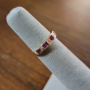 May include: A gold band ring with a row of square-shaped pink gemstones and smaller clear gemstones.