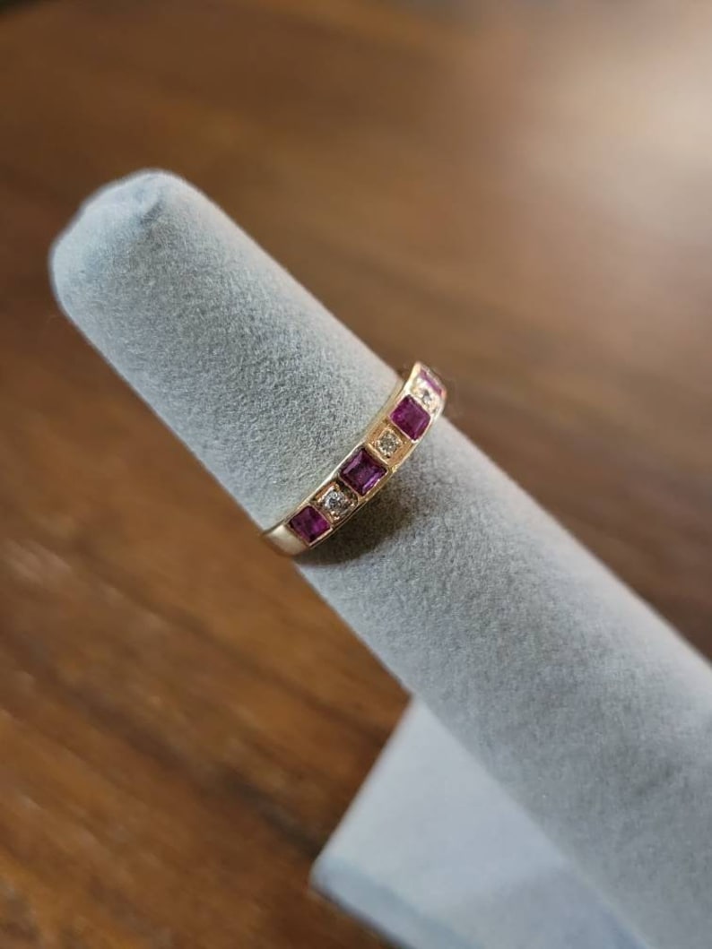 May include: A gold band ring with a row of square-shaped pink gemstones and smaller clear gemstones.