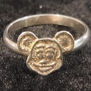 Pu&ograve; includere: Un anello color argento con un ornamento centrale a forma di testa di topo dei cartoni animati, con orecchie rotonde e un volto sorridente. La fascia &egrave; liscia e circolare, mentre il design della testa presenta una superficie ruvida e granulosa.