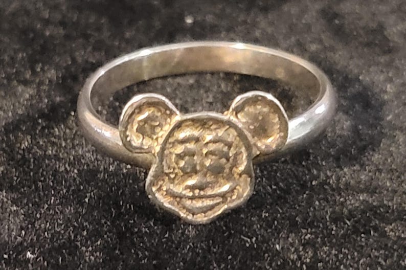 Pu&ograve; includere: Un anello color argento con un ornamento centrale a forma di testa di topo dei cartoni animati, con orecchie rotonde e un volto sorridente. La fascia &egrave; liscia e circolare, mentre il design della testa presenta una superficie ruvida e granulosa.