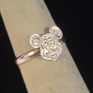 Pu&ograve; includere: Un anello color argento con la testa scolpita e tridimensionale di un topo dei cartoni animati, caratterizzato da grandi orecchie arrotondate, un'ampia bocca sorridente e tratti del viso definiti. L'anello in metallo &egrave; liscio e lucido.