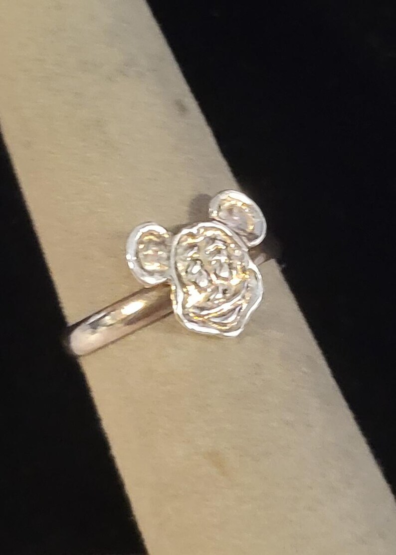 Pu&ograve; includere: Un anello color argento con la testa scolpita e tridimensionale di un topo dei cartoni animati, caratterizzato da grandi orecchie arrotondate, un'ampia bocca sorridente e tratti del viso definiti. L'anello in metallo &egrave; liscio e lucido.