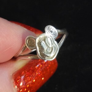 Puede incluir: Un anillo de tono plateado que presenta una cabeza de personaje estilizada en relieve con dos orejas redondeadas. El anillo tiene un dise&ntilde;o de banda dividida que se conecta al motivo central. La superficie met&aacute;lica tiene un acabado pulido.